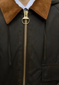 Veste noire avec col en velours côtelé marron et grande tirette de fermeture éclair ronde en laiton sur col de chemise blanc et cravate bleue.