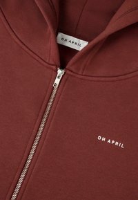 Felpa con zip color marrone bordeaux realizzata in morbido tessuto, con una zip argentata e un logo bianco "OH APRIL" ricamato sul petto. Texture liscia.