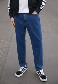 Blue denim straight-leg jeans with a smooth texture, worn with a black zip-up jacket and black-and-white sneakers.