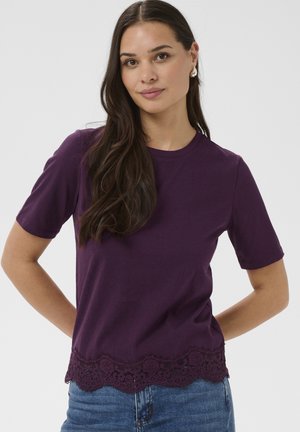 Kobieta o długich brązowych włosach, ubrana w fioletowy krótki top z koronkowym wykończeniem i niebieskie dżinsy, stojąca z rękami za plecami.