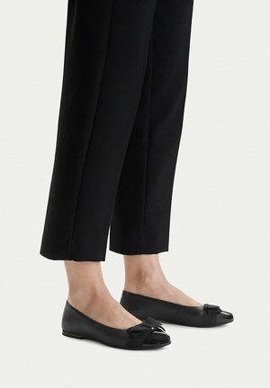 Pies con zapatos planos negros con detalle de lazo, combinados con pantalones negros cortos sobre un fondo blanco.