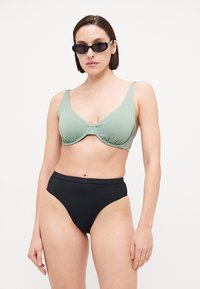 Top bikini verde a coste con ferretto e dettaglio annodato, abbinato a slip bikini neri a vita alta. Modella che indossa occhiali da sole neri.
