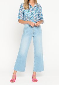 Camicia blu a fantasia con maniche corte, jeans azzurri a gamba larga e sandali rosa con cinturini. Presenti trame e contrasti di colore notevoli.