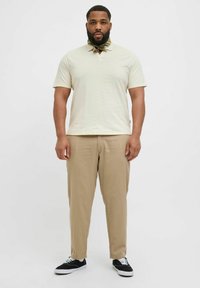 Světle béžová polo košile s jemnou strukturou, spárovaná s hnědými kalhotami. Černé tenisky s bílými podrážkami. Neutrální barevná paleta po celém outfitu.