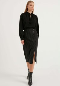 Svart satinstik blus med samlad halsringning, i kombination med en knälång svart kostymkjol med smala ränder, sidoslits och knappdetalj. Svarta klackstövlar.