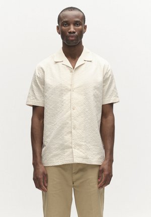 Muž oblečený v krémovobielej krátkoročnej textúrovanej košeli na gombíky a khaki nohaviciach, stojaci pred jednoduchým bielym pozadím.