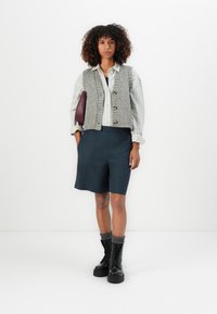 Vrouw met krullend haar, gekleed in een gestreepte shirt, een grijze gebreide vest, donkere korte broek, zwarte combatboots met grijze sokken, en houdt een bordeauxrode clutch vast.