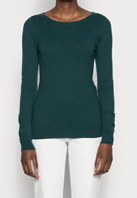 Personne portant un pull ajusté à manches longues vert foncé et un pantalon blanc, se tenant contre un fond clair uni.