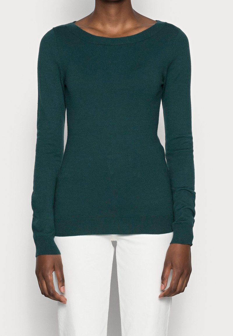 Personne portant un pull ajusté à manches longues vert foncé et un pantalon blanc, se tenant contre un fond clair uni.