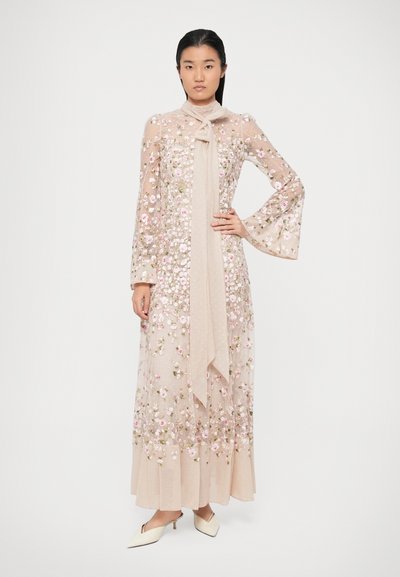 Γυναίκα στέκεται φορώντας ένα μακρύ διαφανές μπεζ φόρεμα με λουλουδάτη κεντητική, φουσκωτά μανίκια, κόμπο στον λαιμό και κρεμ χαμηλοτάκουνες μυτερές γόβες.