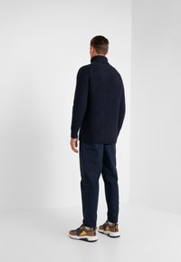 Homme tourné de dos, portant un pull en grosse maille bleu marine, un pantalon foncé et des chaussures de sport marron, debout dans un coin d'une pièce blanche.