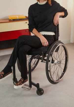 Persoon in zwarte kleding zittend in een handbewogen rolstoel binnenshuis, met één arm rustend op de duwrand van de rolstoel.