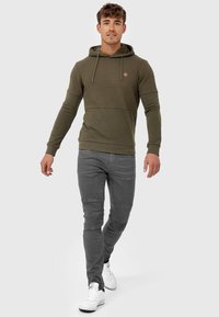 Olijfgroene hoodie met een gestructureerde bovenkant, een trekkoordcapuchon en geribde mouwen, gecombineerd met grijze slim-fit jeans en witte sneakers.