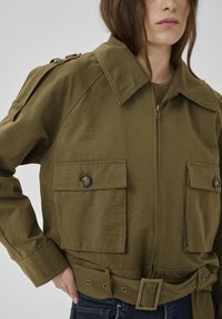 Frau trägt eine olivgrüne Utility-Jacke mit großen, mit Knöpfen verschlossenen Taschen, Reißverschluss vorne, weitem Kragen und tailliertem Gürtel, Hand in der Hüfte.