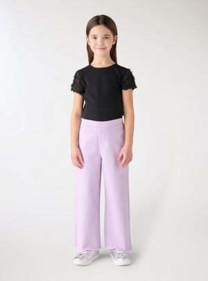 Młoda dziewczyna stoi, ubrana w czarną koszulkę z koronkowymi rękawami, lawendowe szerokie spodnie i srebrne trampki na jednolitym białym tle.