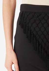 Falda negra con un diseño tejido tipo red que incluye flecos colgantes en un lado y una cintura ajustada. Textura suave con un acabado entallado.