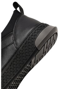 Zapatilla negra con parte superior de cuero suave, diseño flexible, suela de goma texturizada con un patrón geométrico y un asa para facilitar el calzado.