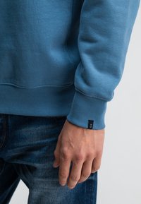 Lichtblauwe sweatshirt met geribbelde manchetten, een ronde halslijn en een klein zwart labeldetail. Draagt met blauwe denim jeans, wat een casual stijl laat zien.