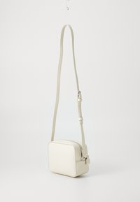 Krämfärgad lädercrossbodyväska med rektangulär form, texturerad yta, justerbar rem och dragkedjestängning med en liten hårdvarudetalj.