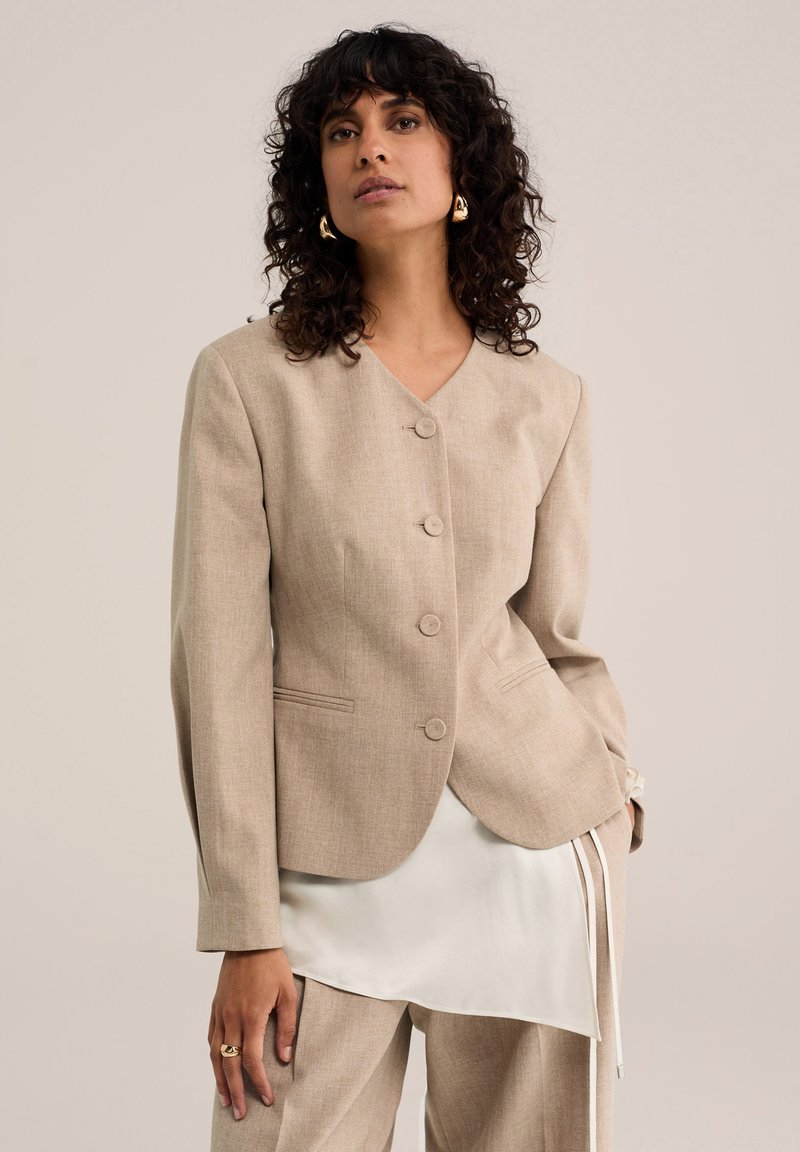 Femme aux cheveux bouclés foncés portant un blazer beige boutonné, un pantalon assorti, un haut blanc superposé, des boucles d’oreilles dorées et une bague, posant devant un fond uni.
