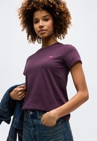 Νεαρή γυναίκα με σγουρά μαλλιά, φορώντας ένα μοβ t-shirt Levi's, μπλε τζιν και κρατώντας ένα τζιν μπουφάν στον ώμο της.