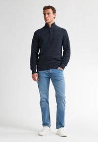 Marineblauwe textuur pullover met een kwart rits, gecombineerd met lichtblauwe jeans en witte sneakers. Eenvoudig ontwerp, minimale branding op de mouw.