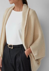 Cardigan en tricot beige à col ouvert, porté sur une chemise blanche, associé à un pantalon noir à rayures et une boucle de ceinture en forme de cœur en or.