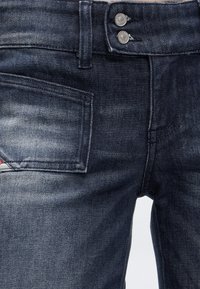Gros plan sur un jean en denim bleu marine avec des boutons argentés doubles et une petite poche montrant une étiquette partiellement visible.