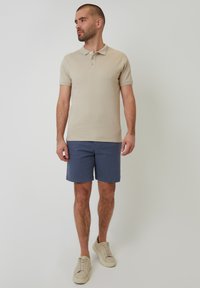 Béžová polo košile s krátkým rukávem a strukturovanou tkaninou, doplněná o námořnické šortky. Lehká obuv dokončuje ležérní outfit.