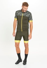 Pyöräilypaita oliivinvihreänä, jossa on harmaa kuvioitu yläosa, keltaiset korostukset, etuvetoketju ja siihen sopivat bib-shortsit keltaisilla nauhoilla. Mustat sukat ja kengät.