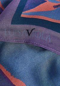 Pañuelo de seda con patrones geométricos en azul, morado y naranja. Bordes acabados cuidadosamente con costuras. Nombre de la marca visible.