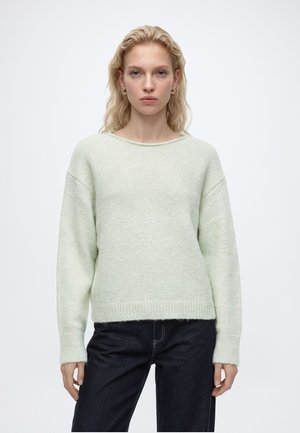 Jeune femme aux cheveux blonds portant un pull en maille vert clair à manches longues et un pantalon foncé, debout devant un fond blanc uni.