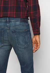 Uomo che indossa jeans in denim blu di Calvin Klein e una camicia a scacchi rossa e nera, mostrato dalla vita in su con la mano destra rilassata.