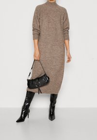 Robe longue en maille marron à manches longues avec un col montant, associée à des bottes noires brillantes montantes jusqu'aux genoux et un petit sac à main noir orné de détails métalliques.