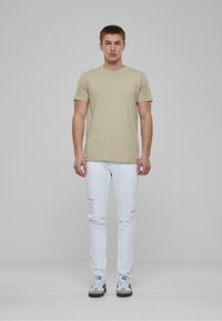 Camiseta de algodón beige, de manga corta y cuello redondo; combinada con jeans blancos desgastados y zapatillas negras/blancas con acentos azules. Diseño sencillo.