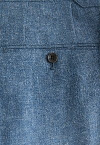 Denim-Stoff in einem mittleren Blauton, mit einem einzelnen schwarzen Knopf und einem subtilen strukturierten Muster auf der Oberfläche.