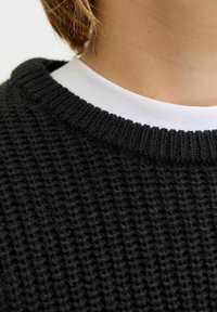 Schwarzer gestrickter Pullover mit einem strukturierten Muster, der über einem weißen Kragenhemd mit geripptem Rundhalsausschnitt getragen wird.