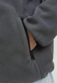 Giacca in pile grigia con una morbida texture, dotata di una tasca laterale con zip e una vestibilità rilassata. Il tessuto è caldo e leggero.