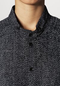 Chemise noire avec un motif texturé de points blancs, dotée d'un col classique et de boutons noirs le long de la patte de boutonnage avant.