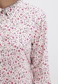 Blomstrig skjorta med knappar i vitt med rosa och lila blommönster. Har en klassisk krage och knäppningar fram.