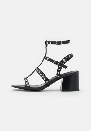 Schwarze Sandale mit Absatz und Nieten, ausgestattet mit einem T-Riemen-Design und verstellbarer Schnalle am Knöchel. Hergestellt aus strukturiertem Material, mit einem Blockabsatz.