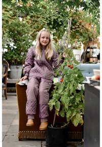 Quilted mauve outdoor outfit on a child, sitting on a brown cushion, holding a tomato plant with red tomatoes. Natural setting, green foliage.
