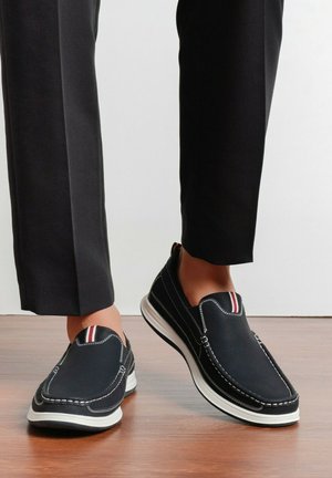 Chaussures décontractées noires à enfiler avec des coutures blanches et une semelle blanche, portées avec un pantalon noir sur un sol en bois.