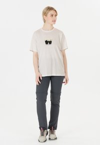 Ljusbeige t-shirt med en berggrafik, kombinerad med mörkgrå byxor. Modell i beige vandringsskor, stående mot en enfärgad bakgrund.