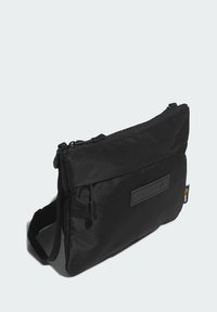 Borsa a tracolla nera realizzata in tessuto resistente, dotata di chiusura con cerniera, tracolla regolabile e una toppa testurizzata con branding sul davanti.