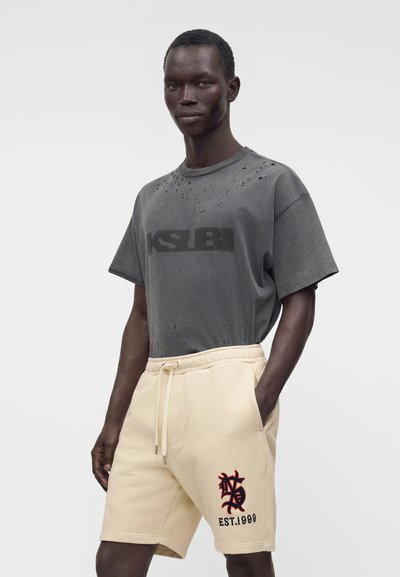 Homme à la peau foncée portant un t-shirt gris foncé usé et un short beige à cordon avec un emblème noir et rouge ainsi que le texte EST. 1999.