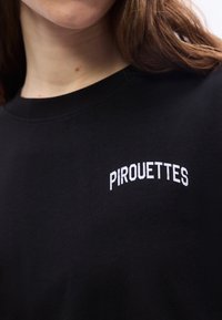 Κοντινό πλάνο ενός ατόμου που φοράει μαύρο πουκάμισο με λευκό κείμενο "PIROUETTES" τυπωμένο στην περιοχή του στήθους.