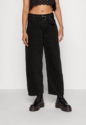 Person wearing high-waisted wide-leg black jeans with buckle belt, black lace-up platform boots, and cropped black top against white background.