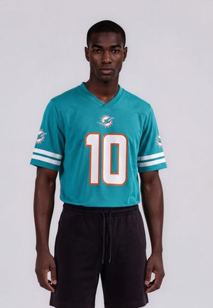 Man die een teal Miami Dolphins footballshirt met nummer 10 draagt en zwarte shorts, staat voor een effen lichte achtergrond.