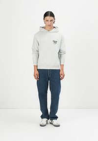 Grå hettegenser med en svart logo på brystet, kombinert med blå jeans og hvite joggesko med svarte striper. Enkel bakgrunn.